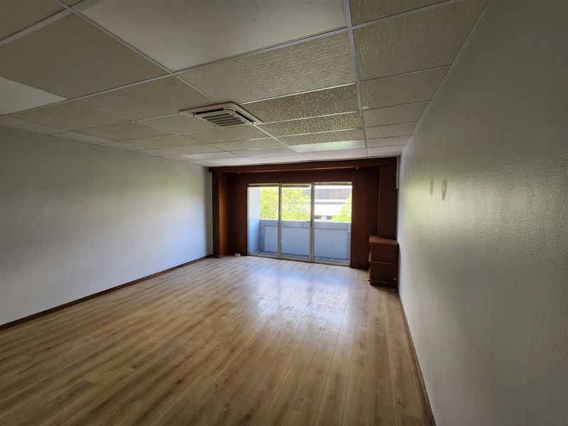 An empty office space featuring laminate flooring, a suspended ceiling with integrated air conditioning, and a sliding glass door leading to a balcony.