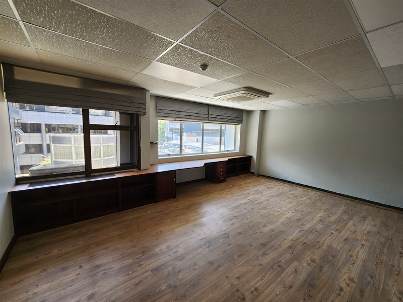 A spacious, empty office featuring wood-look laminate flooring, built-in cabinetry along the windows, a suspended ceiling, and a cassette air conditioning unit.