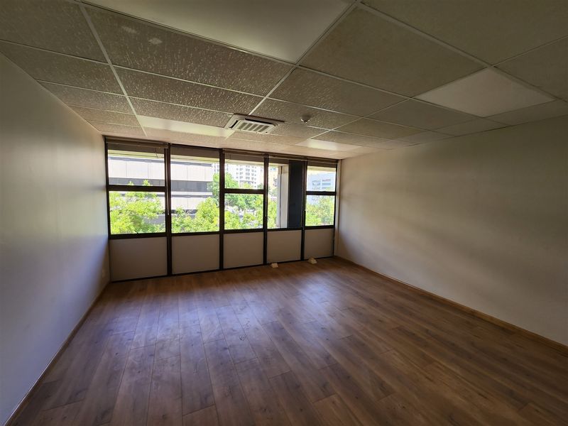An empty office space featuring laminate flooring, a suspended ceiling with integrated air conditioning, and large windows providing natural light.