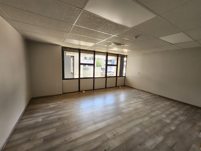 A bright, empty office space featuring modern laminate flooring, a suspended ceiling with recessed lighting, and large windows providing natural light.