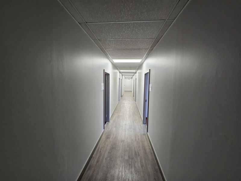 A clean, well-lit office hallway featuring modern laminate flooring, a suspended ceiling with recessed lighting, and multiple access doors.