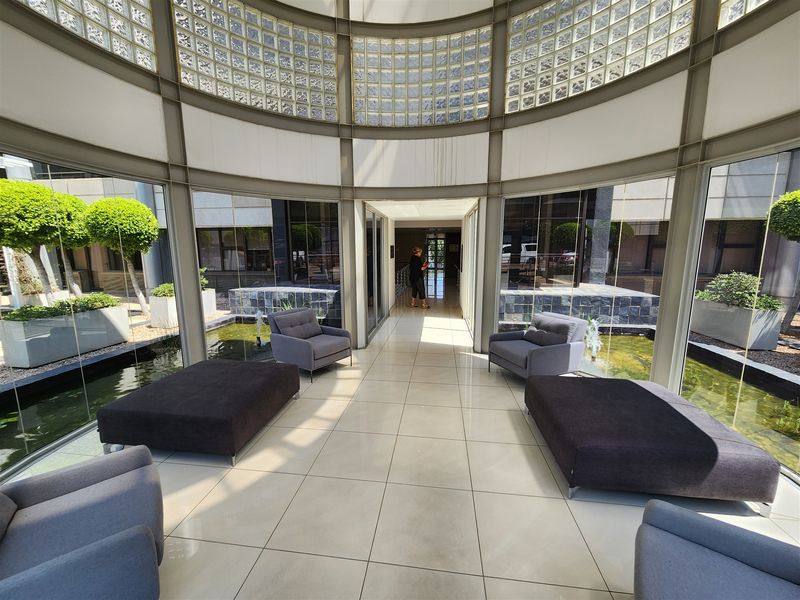 A bright, modern office entrance lobby featuring floor-to-ceiling glass walls, tiled flooring, and a central water feature under a glass block clerestory.