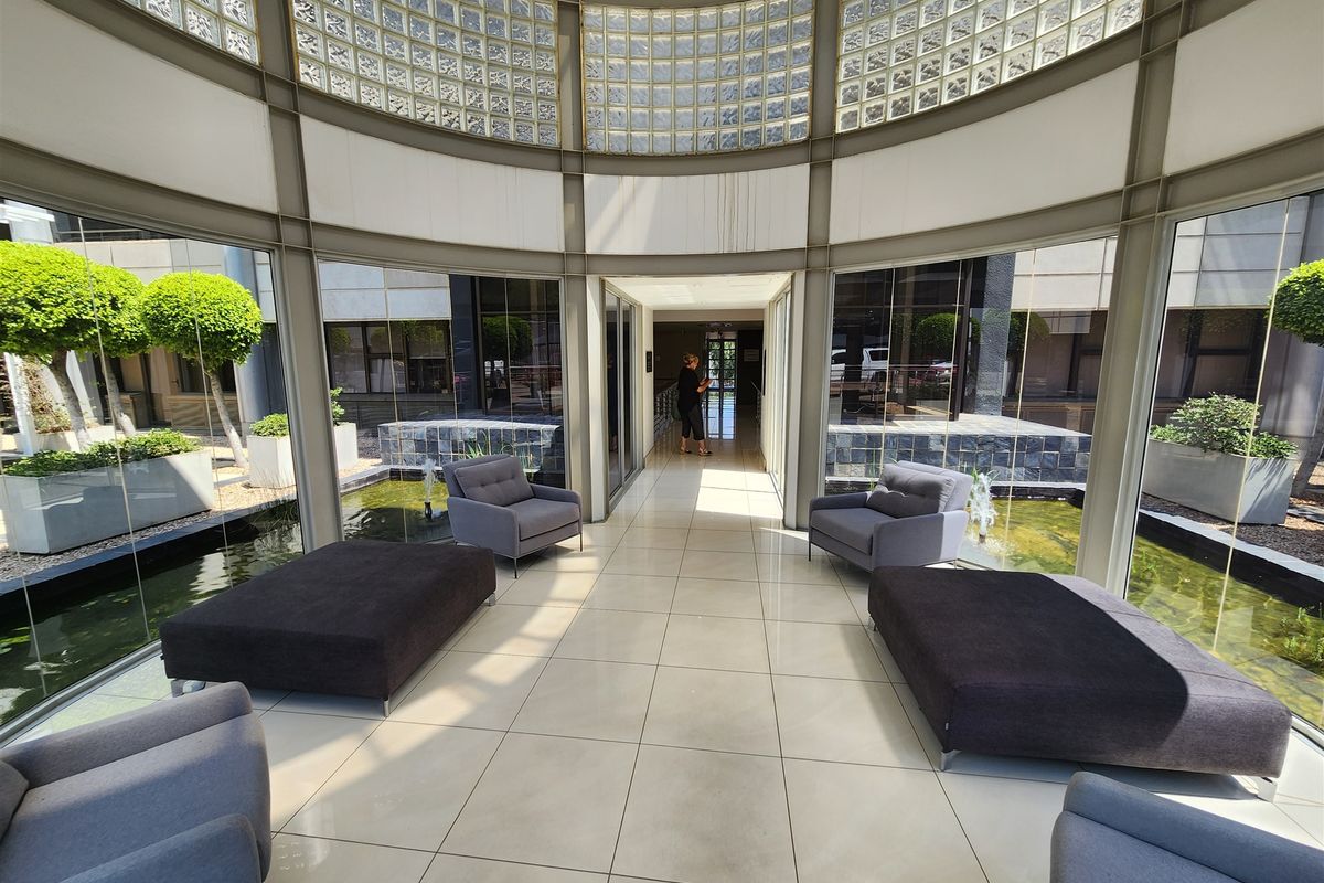 A bright, modern office entrance lobby featuring floor-to-ceiling glass walls, tiled flooring, and a central water feature under a glass block clerestory.