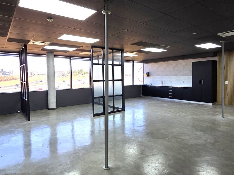 Modern open-plan office space featuring polished concrete floors, black framed glass partitions, a sleek kitchenette, and expansive windows providing ample natural light.