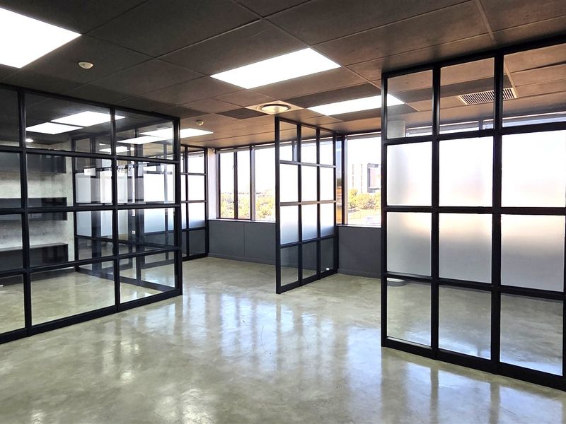 Modern open-plan office space featuring polished concrete floors, black metal-framed glass partitions, and large windows providing ample natural light.