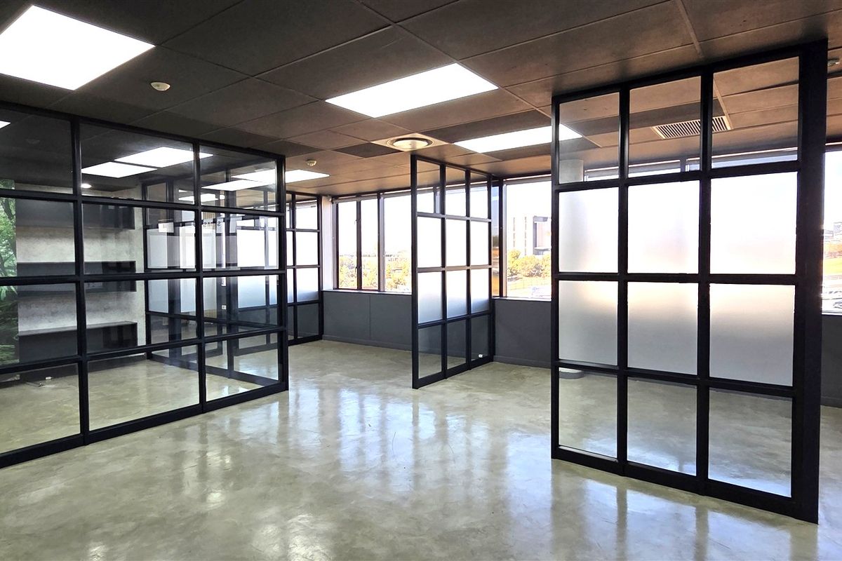 Modern open-plan office space featuring polished concrete floors, black metal-framed glass partitions, and large windows providing ample natural light.