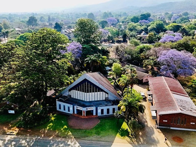 Aerial view of a commercial property complex featuring a prominent pitched-roof building, surrounding landscaped grounds, and paved access areas in a lush setting.