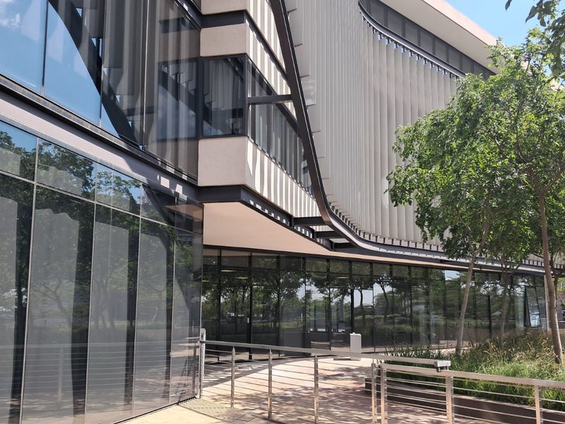 Modern office building exterior featuring extensive glass curtain walling, architectural louvers, and a paved walkway with stainless steel railings.