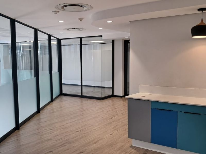 Modern office space featuring glass-partitioned meeting rooms, wood-look flooring, recessed ceiling lighting, and a compact kitchenette area with built-in cabinetry.
