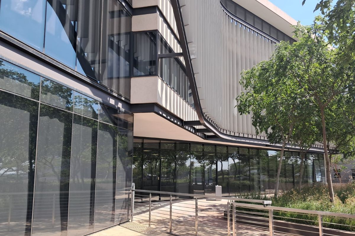 Modern office building exterior featuring extensive glass curtain walling, architectural louvers, and a paved walkway with stainless steel railings.