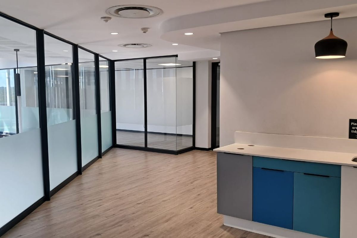 Modern office space featuring glass-partitioned meeting rooms, wood-look flooring, recessed ceiling lighting, and a compact kitchenette area with built-in cabinetry.