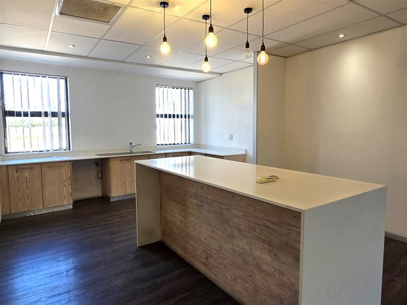 A clean, modern office kitchen area featuring a central island, light wood cabinetry, stone countertops, and a suspended ceiling with pendant lighting.