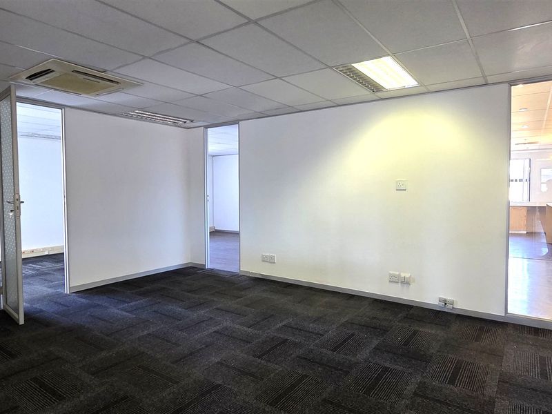 A bright, open-plan office space featuring dark carpet tiles, a suspended ceiling with recessed lighting, and a glass partition wall.