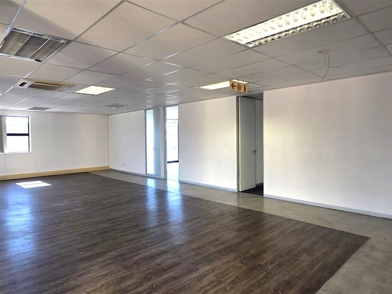 A spacious, vacant open-plan office area featuring a mix of laminate and concrete flooring, suspended ceilings with recessed lighting, and central air conditioning.
