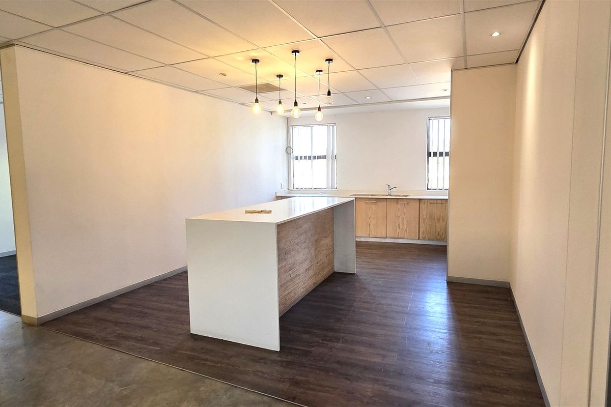 A clean, modern office kitchenette featuring a central island, built-in cabinetry with a sink, and a suspended ceiling with integrated lighting.