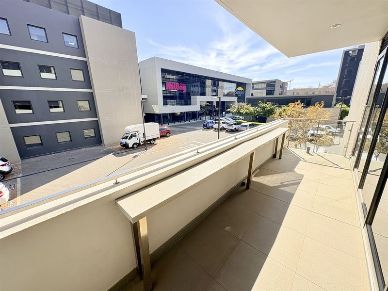 A modern, tiled balcony featuring a stainless steel balustrade and a built-in counter, overlooking a commercial office park area.