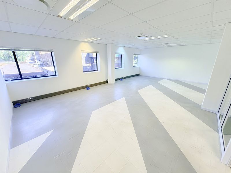 A bright, vacant open-plan office space featuring a suspended ceiling, recessed lighting, and patterned vinyl flooring with large windows providing natural light.