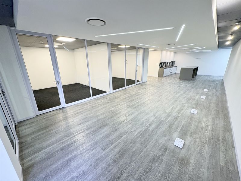 Modern open-plan office space featuring glass-partitioned private offices, sleek laminate flooring, recessed linear lighting, and a compact built-in kitchenette area.