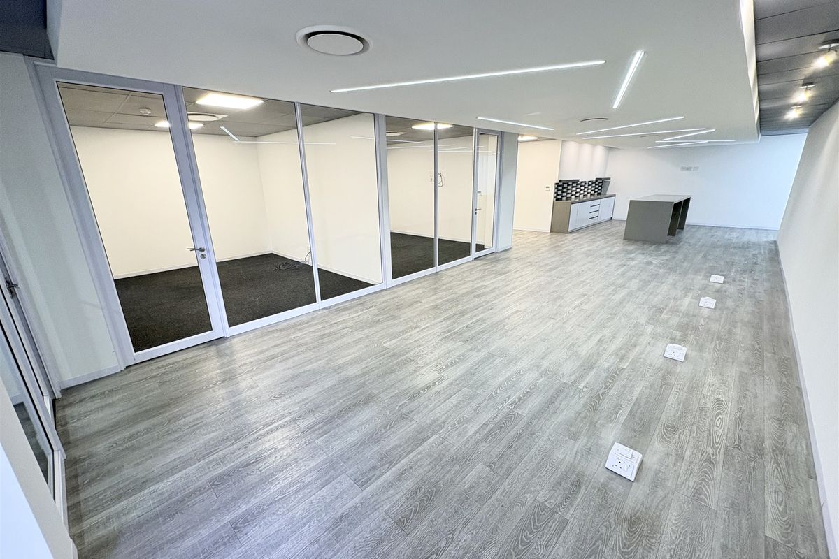 Modern open-plan office space featuring glass-partitioned private offices, sleek laminate flooring, recessed linear lighting, and a compact built-in kitchenette area.