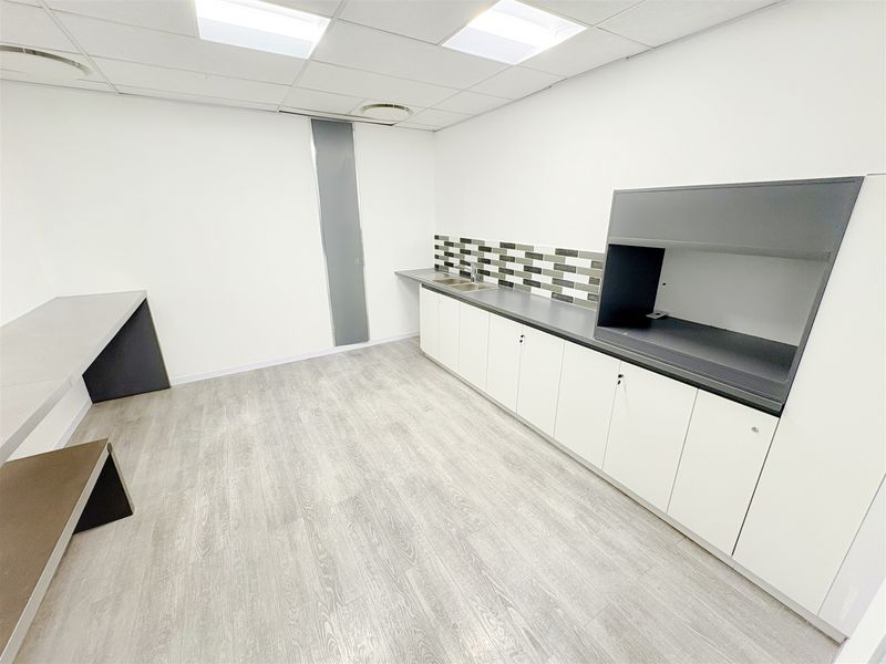 A clean, modern office kitchenette featuring white built-in cabinetry, a dark laminate countertop, stainless steel sink, and a decorative tiled backsplash.