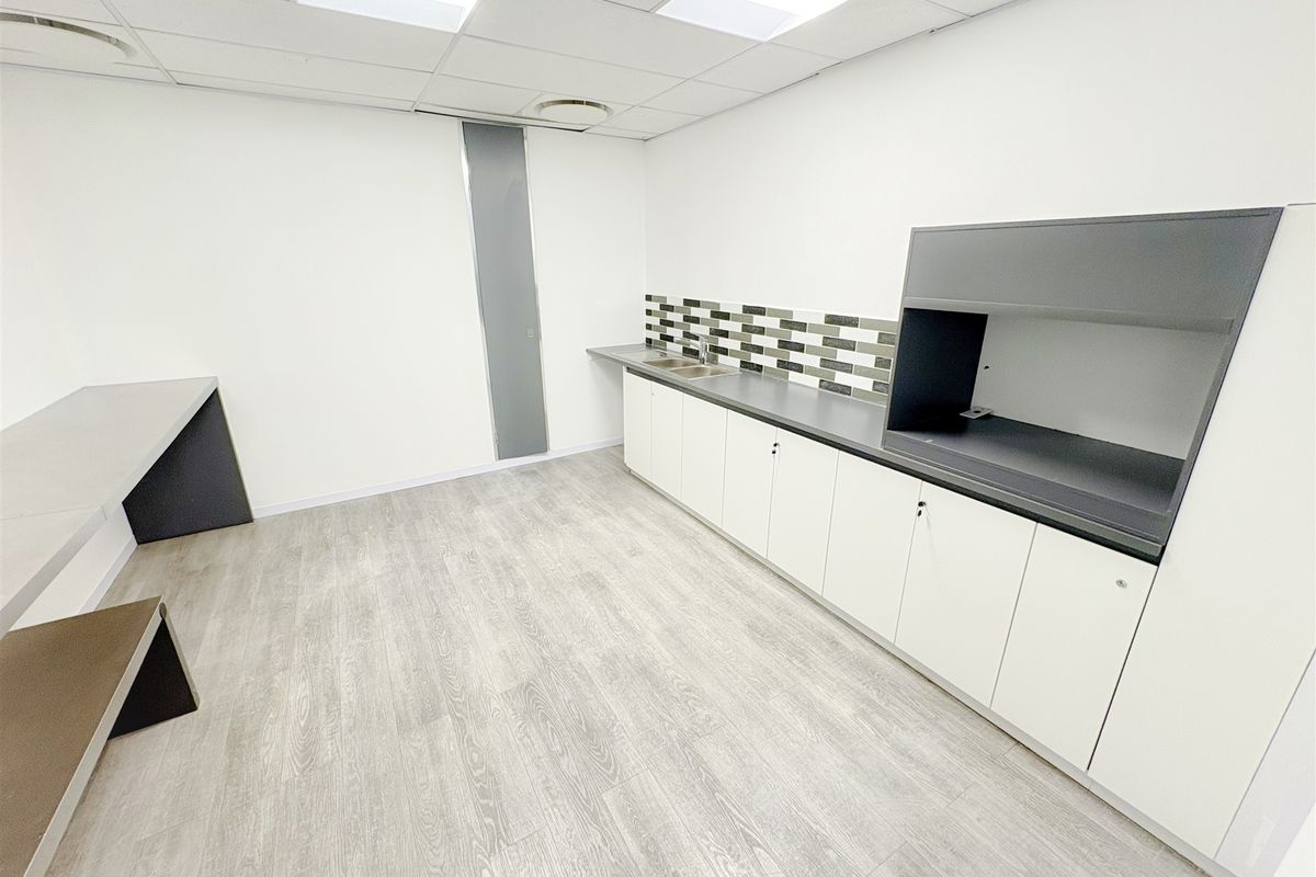 A clean, modern office kitchenette featuring white built-in cabinetry, a dark laminate countertop, stainless steel sink, and a decorative tiled backsplash.