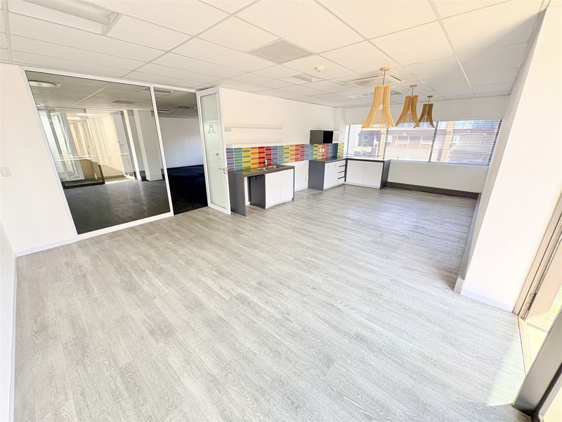 A bright, open-plan office space featuring a modern kitchenette with colorful tiled backsplash, wood-look laminate flooring, and a glass-partitioned meeting area.