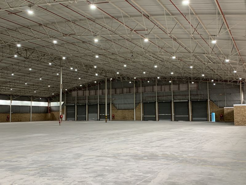 Spacious industrial warehouse featuring high ceilings with exposed steel trusses, multiple roller shutter doors, concrete flooring, and efficient high-bay lighting throughout.