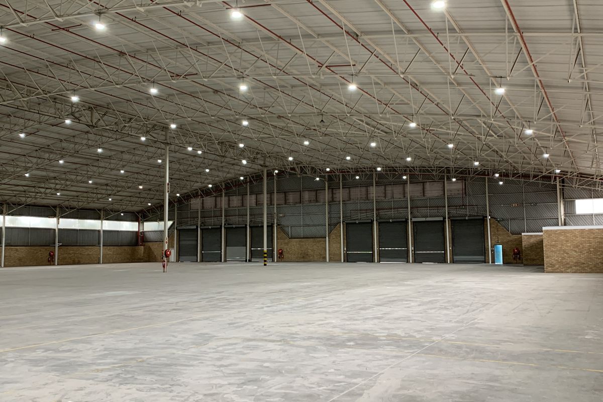 Spacious industrial warehouse featuring high ceilings with exposed steel trusses, multiple roller shutter doors, concrete flooring, and efficient high-bay lighting throughout.