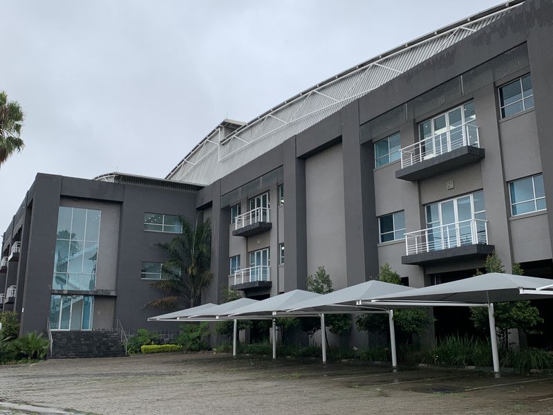 Front elevation of a multi-story industrial office building featuring a paved parking area with shade cloth carports and multiple balconies.