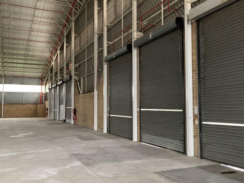 Spacious industrial warehouse interior featuring high ceilings with exposed steel trusses, concrete flooring, and multiple large roller shutter doors for efficient loading access.
