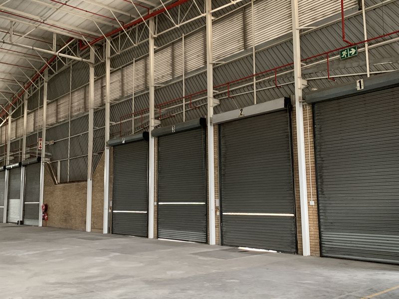 Spacious industrial warehouse interior featuring multiple numbered roller shutter doors, exposed steel roof trusses, and a functional fire sprinkler system.