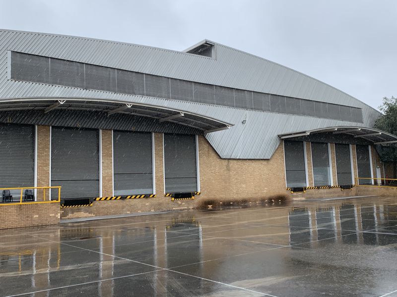 Industrial warehouse exterior featuring multiple roller shutter loading bays, a metal roof canopy, and a spacious paved yard area.