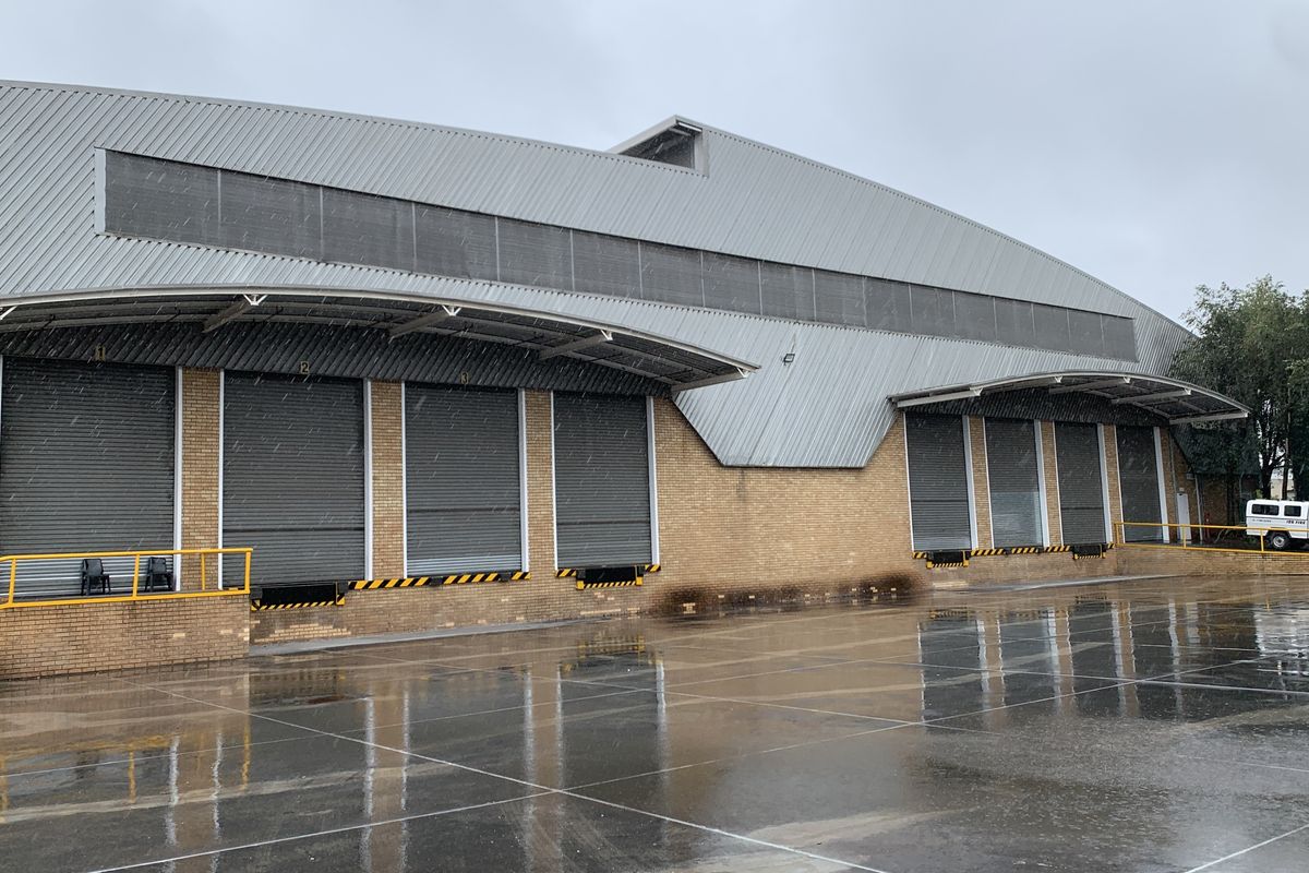 Industrial warehouse exterior featuring multiple roller shutter loading bays, a metal roof canopy, and a spacious paved yard area.