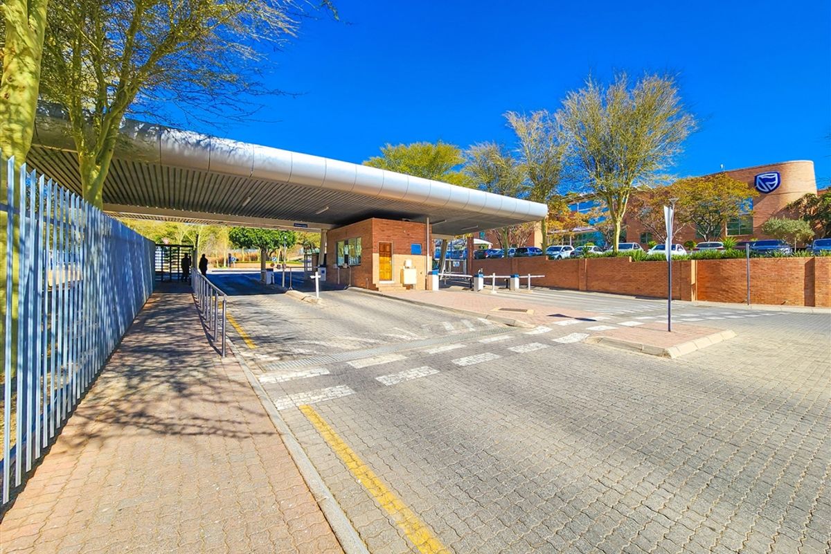 A professional view of the office park entrance featuring a secure boom gate, guard house, and paved driveway access.
