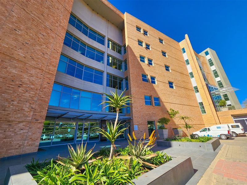 Modern multi-story office building featuring a brick and glass facade, glass entrance, and landscaped exterior planters.