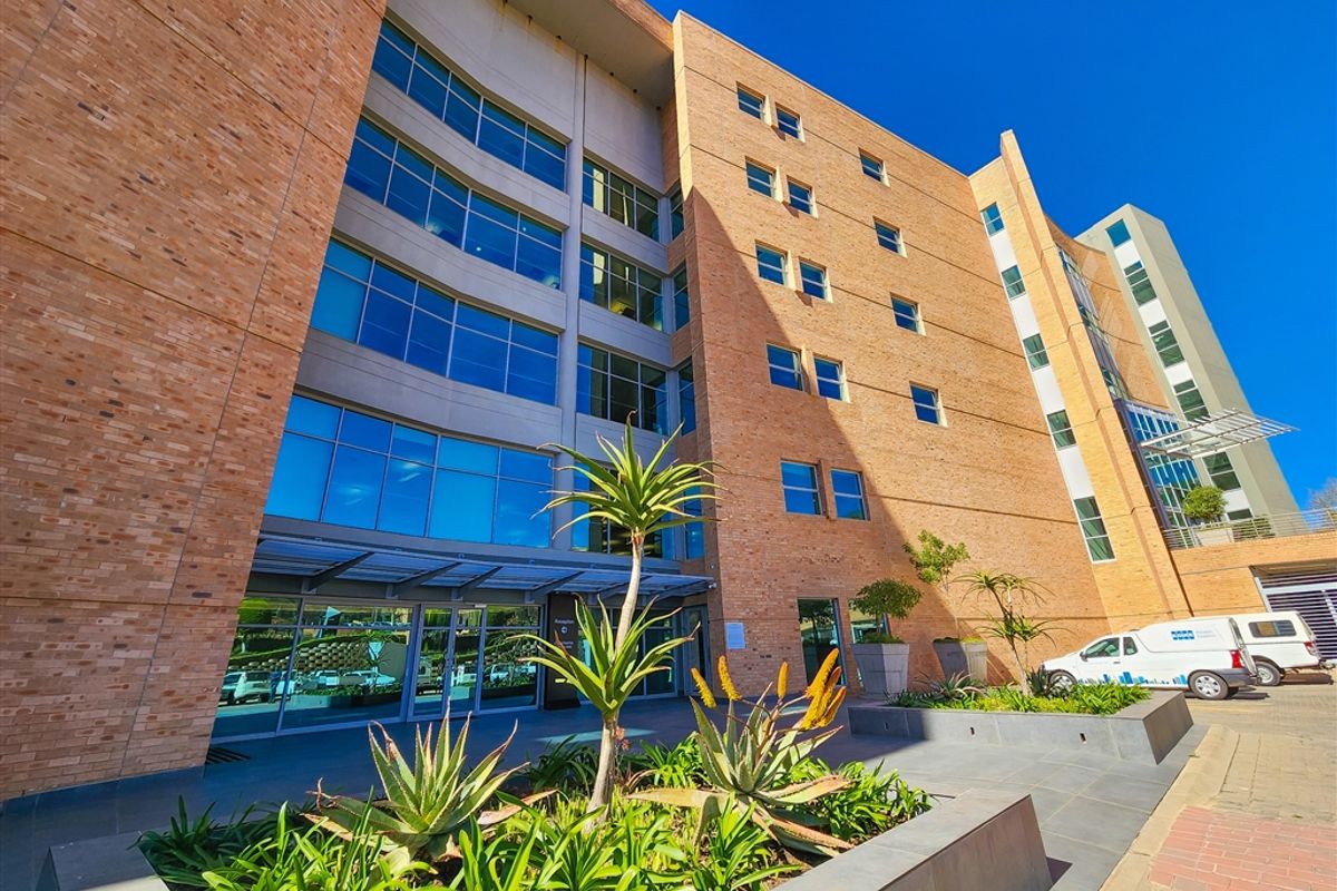 Modern multi-story office building featuring a brick and glass facade, glass entrance, and landscaped exterior planters.