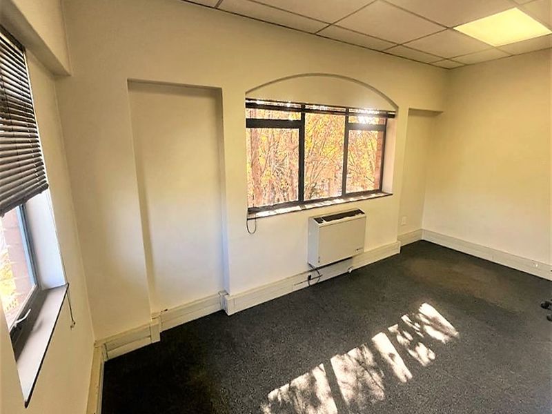 A vacant office space featuring a suspended ceiling, recessed lighting, carpeted flooring, and a wall-mounted HVAC unit with natural light from large windows.