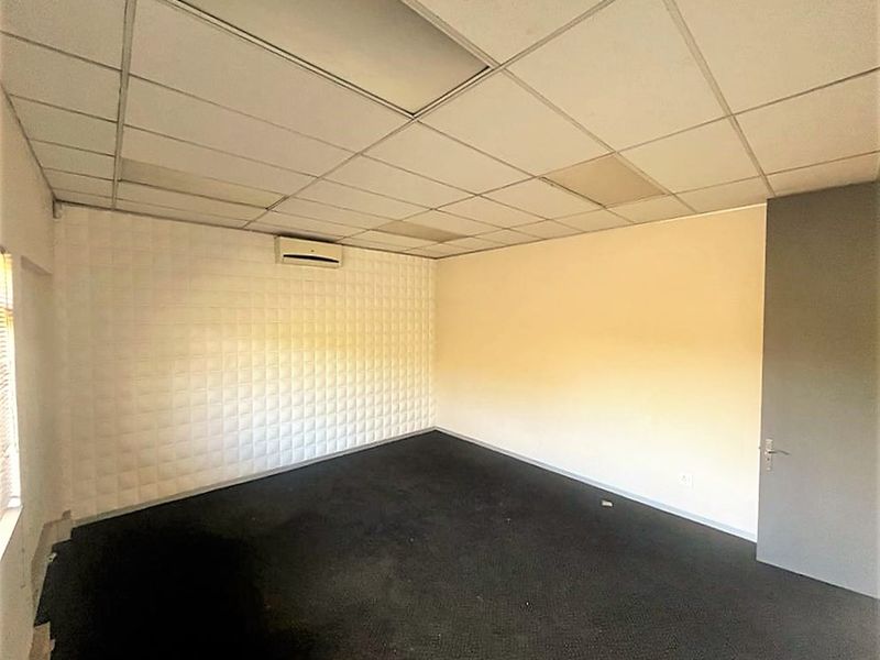 An empty office space featuring a suspended ceiling, wall-mounted air conditioning unit, textured wall paneling, and fitted carpeting.