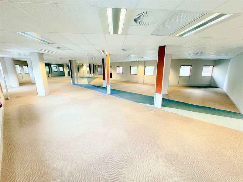 A large, open-plan office space featuring a suspended ceiling with recessed lighting, air conditioning vents, and neutral carpet tiles throughout.