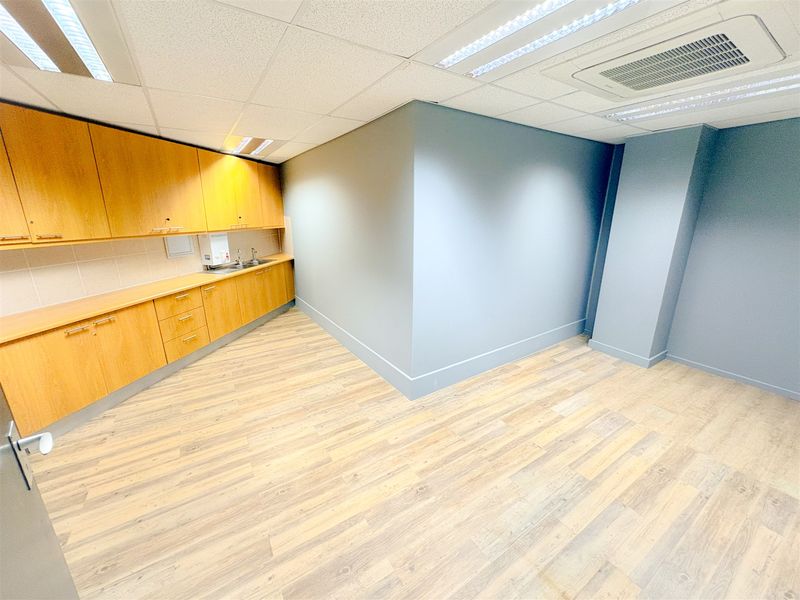 A clean, empty office space featuring wood-look laminate flooring, built-in cabinetry with a sink, and a modern suspended ceiling with integrated lighting and air conditioning.