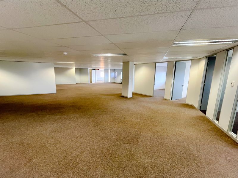 A spacious, open-plan office area featuring neutral carpeting, suspended ceilings with recessed lighting, and glass-partitioned private office spaces.