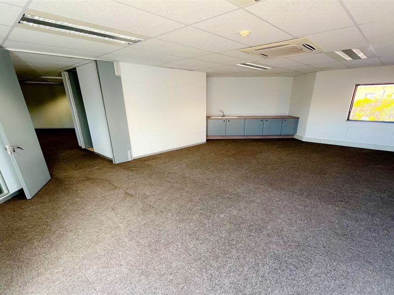 An open-plan office space featuring carpeted flooring, a suspended ceiling with recessed lighting, integrated air conditioning, and a small built-in kitchenette area.