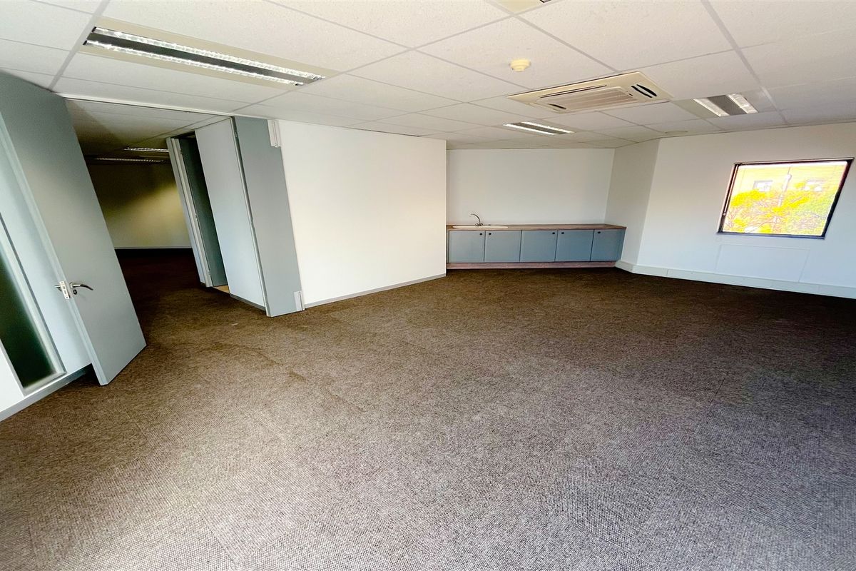 An open-plan office space featuring carpeted flooring, a suspended ceiling with recessed lighting, integrated air conditioning, and a small built-in kitchenette area.