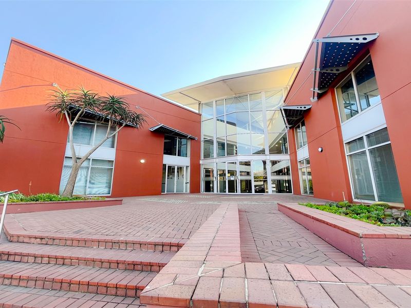 Modern office building entrance featuring a prominent glass facade, brick paved courtyard, and structured metal awnings.