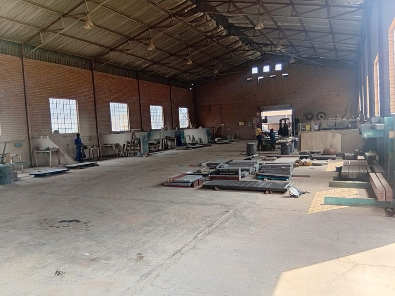Spacious industrial warehouse featuring exposed brick walls, high steel truss ceilings, concrete flooring, and large windows providing ample natural light.
