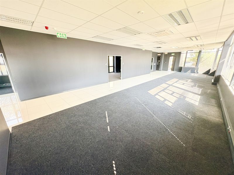 A spacious, vacant open-plan office area featuring a mix of carpet and tiled flooring, suspended ceilings, and large windows providing natural light.