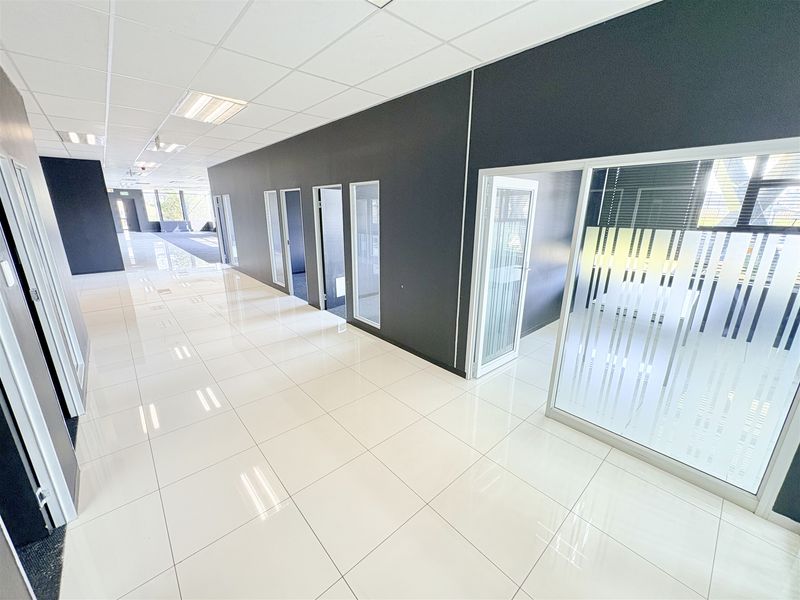 Modern office hallway featuring polished white tiled flooring, suspended ceiling with recessed lighting, and glass-partitioned office suites with frosted privacy detailing.