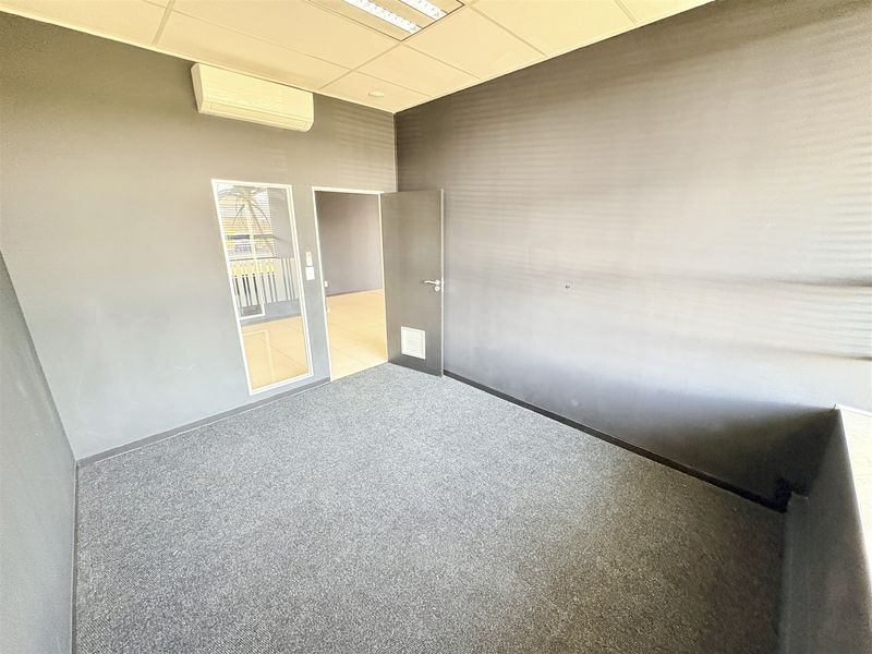 A clean, empty office space featuring grey carpet flooring, a suspended ceiling, and a wall-mounted split-system air conditioner.