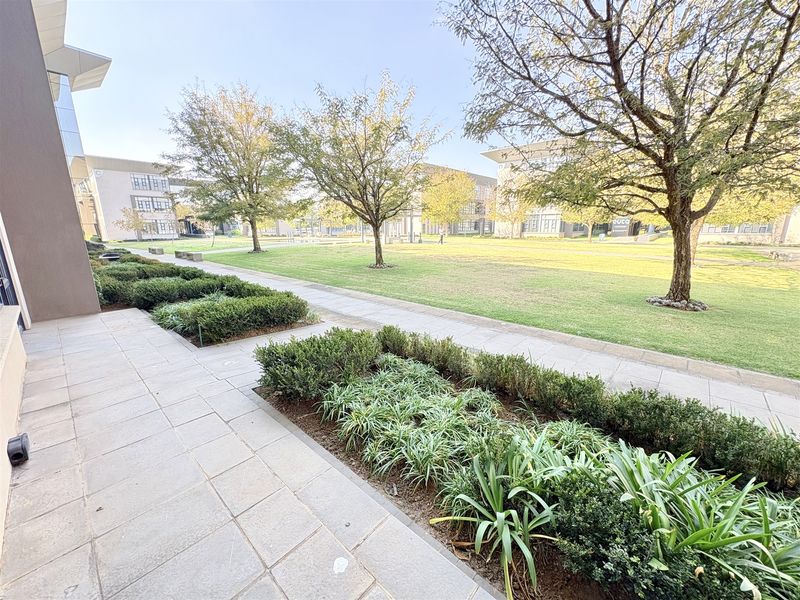 A well-maintained office park exterior featuring a paved walkway, manicured garden beds, and mature trees set against modern office building architecture.