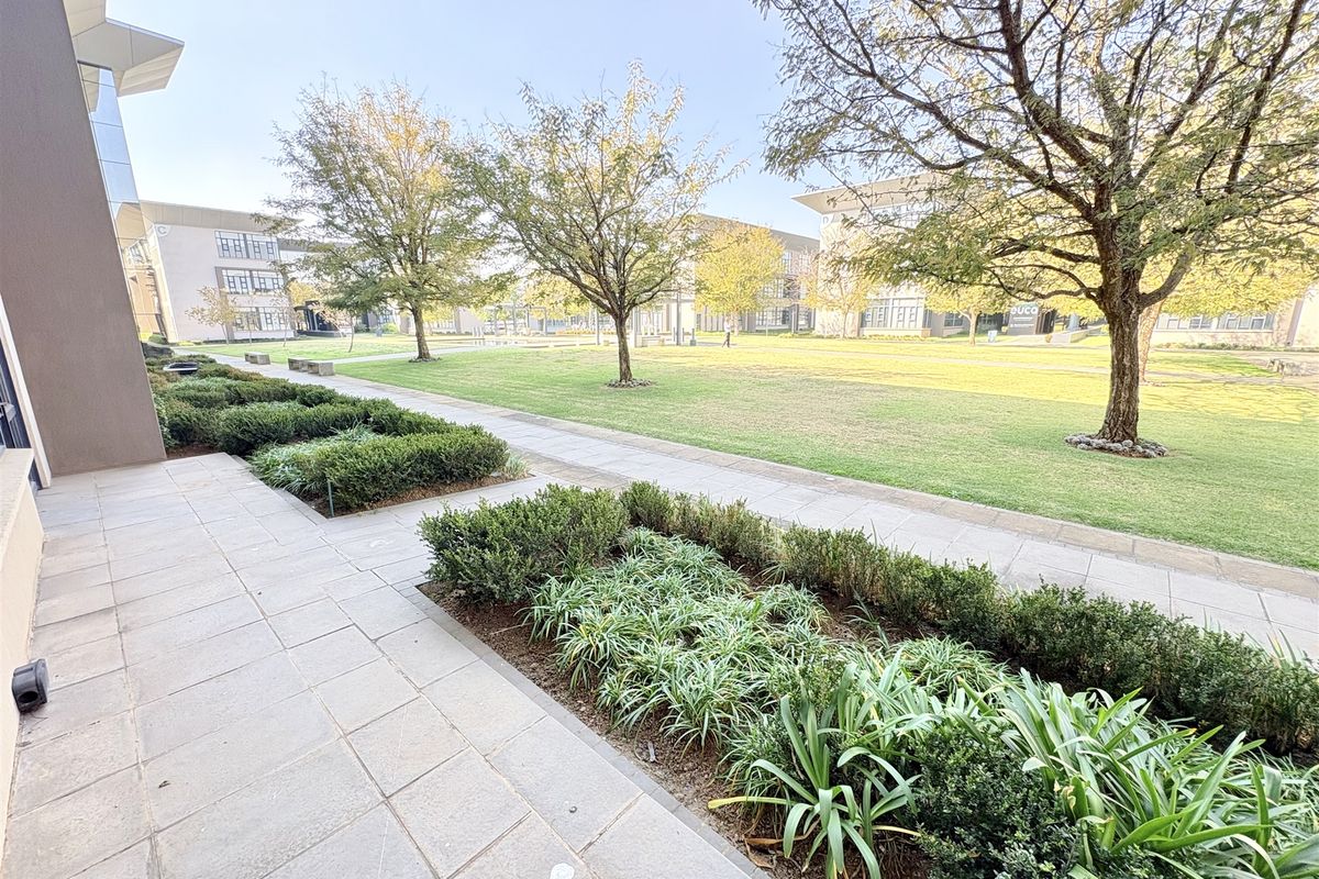 A well-maintained office park exterior featuring a paved walkway, manicured garden beds, and mature trees set against modern office building architecture.